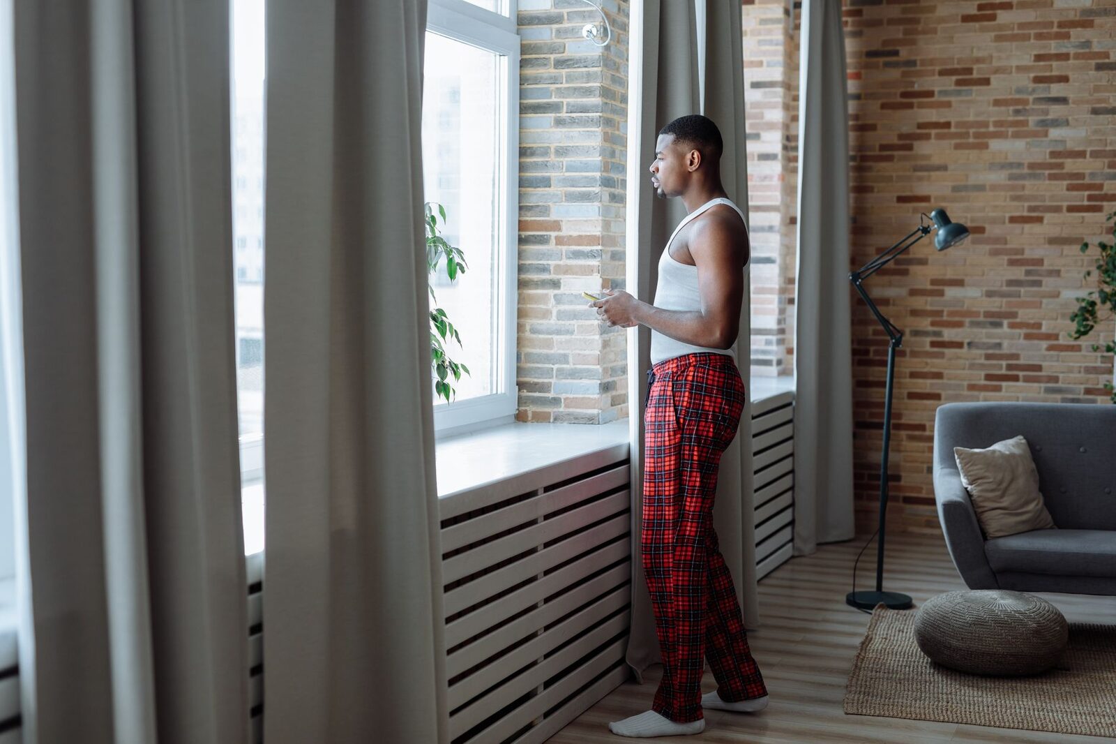 A man in a white tank top and red plaid pants gazes out the window in a modern living room.